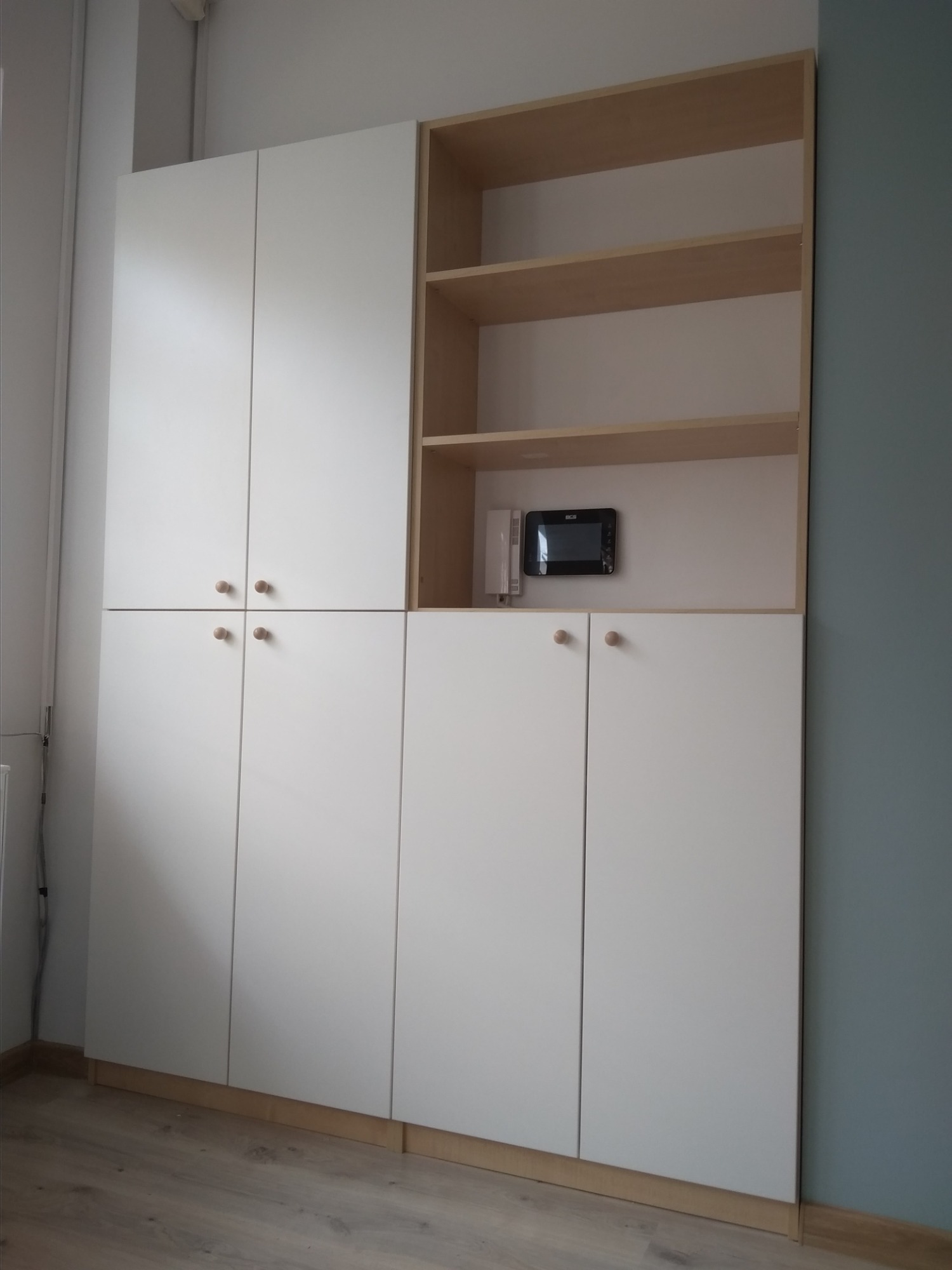 Biała szafa z drewnianymi półkami i okrągłymi uchwytami, z zamontowanym panelem domofonu, na tle ściany w kolorze gołębim.