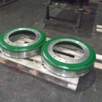 Dwa metalowe pierścienie z zielonymi obudowami na drewnianej palecie w hali produkcyjnej.