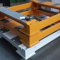 Żółta, stalowa rama konstrukcyjna na białej palecie, widoczne elementy spawane i śruby mocujące, oświetlenie studyjne.