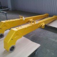 Dwa żółte, metalowe elementy konstrukcyjne, prawdopodobnie dźwigowe, leżące na podłodze w hali produkcyjnej. Widoczne ślady farby i spawania.