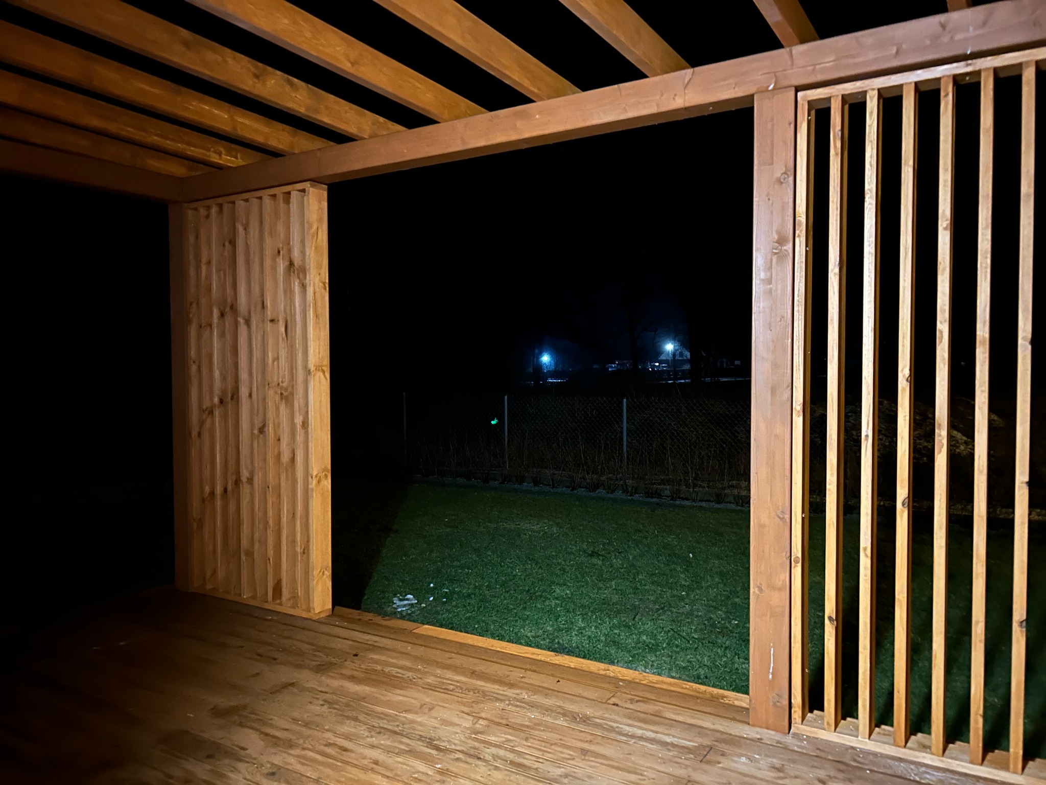 Drewniana pergola z ażurowymi ścianami, oświetlona nocą. Widok na trawnik i oddalone światła. Deski podłogowe w ciepłym odcieniu brązu.