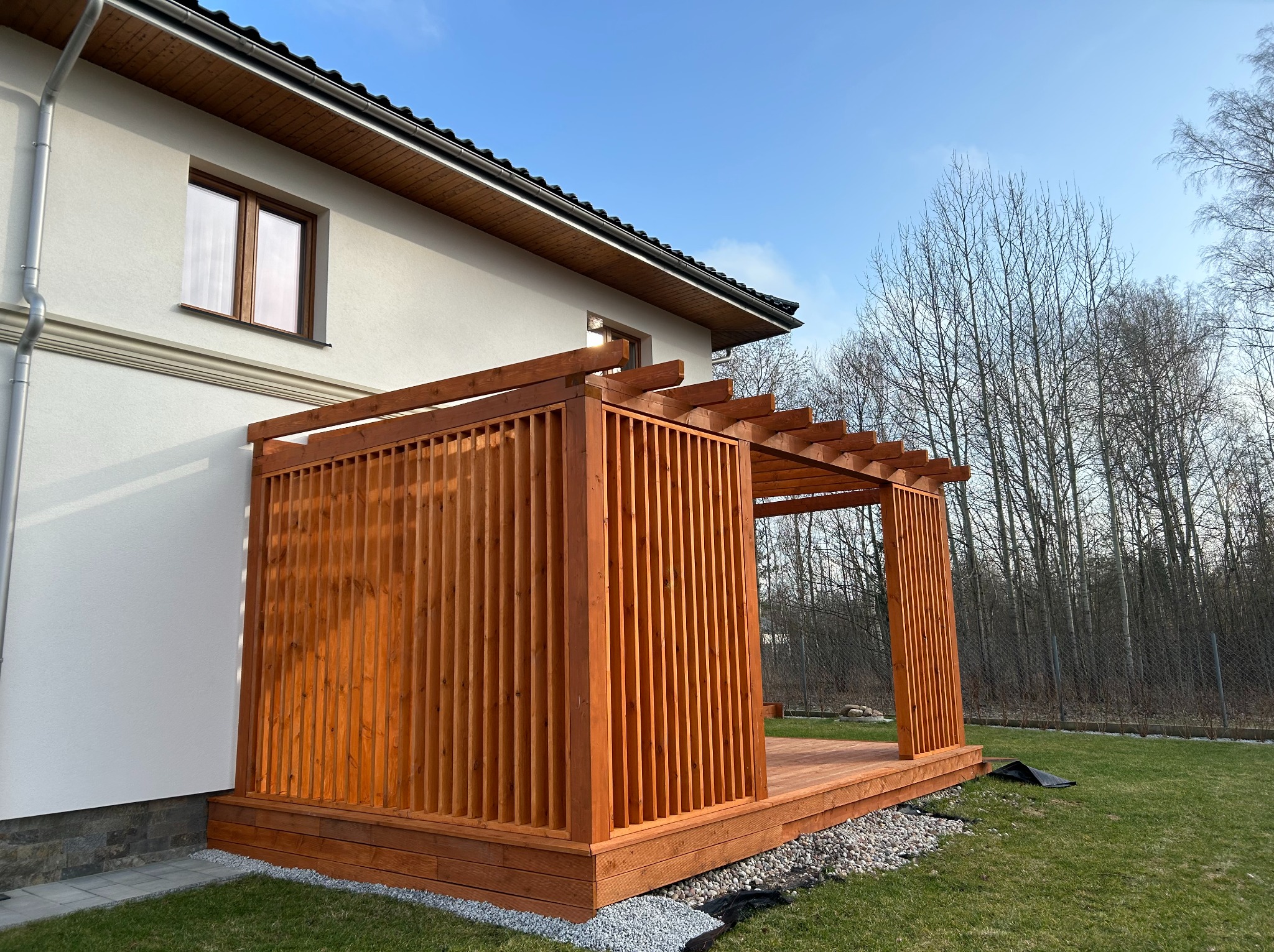 Drewniana pergola z ażurową ścianką przy białym domu, otoczona zielonym trawnikiem i drzewami bez liści, w słoneczny dzień. Widok z boku.