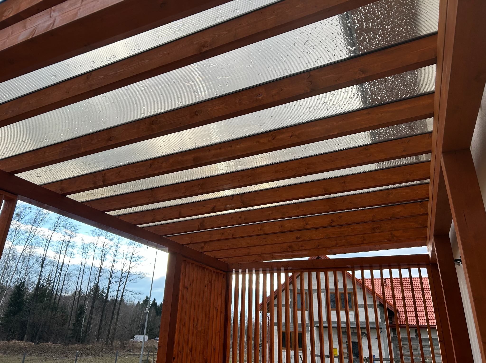 Drewniana pergola z przezroczystym zadaszeniem, widok z dołu. W tle dom i drzewa. Widoczne krople wody na zadaszeniu.