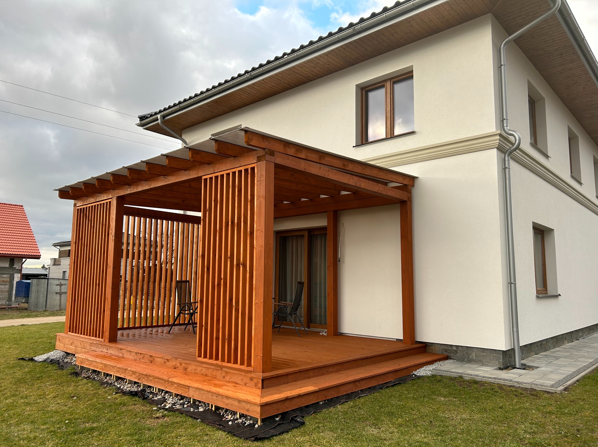 Drewniana pergola z ażurowymi ścianami i schodkowym tarasem przylegająca do białego domu. Widoczne krzesła i elementy wykończenia.
