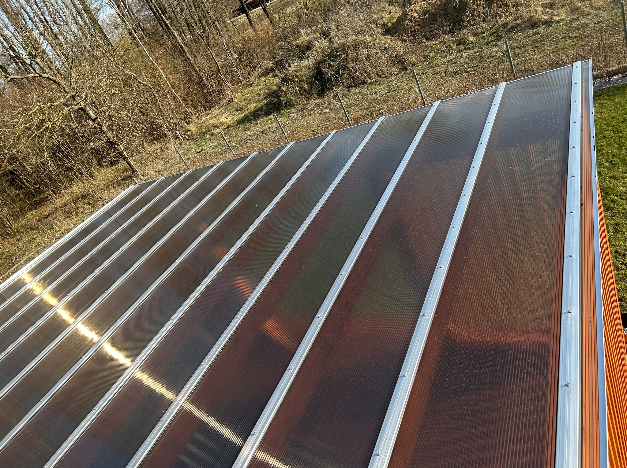 Zadaszenie tarasu z brązowego poliwęglanu i srebrnych profili, widoczne z góry na tle zieleni i drzew. Konstrukcja oświetlona słońcem, z widocznymi detalami mocowań.