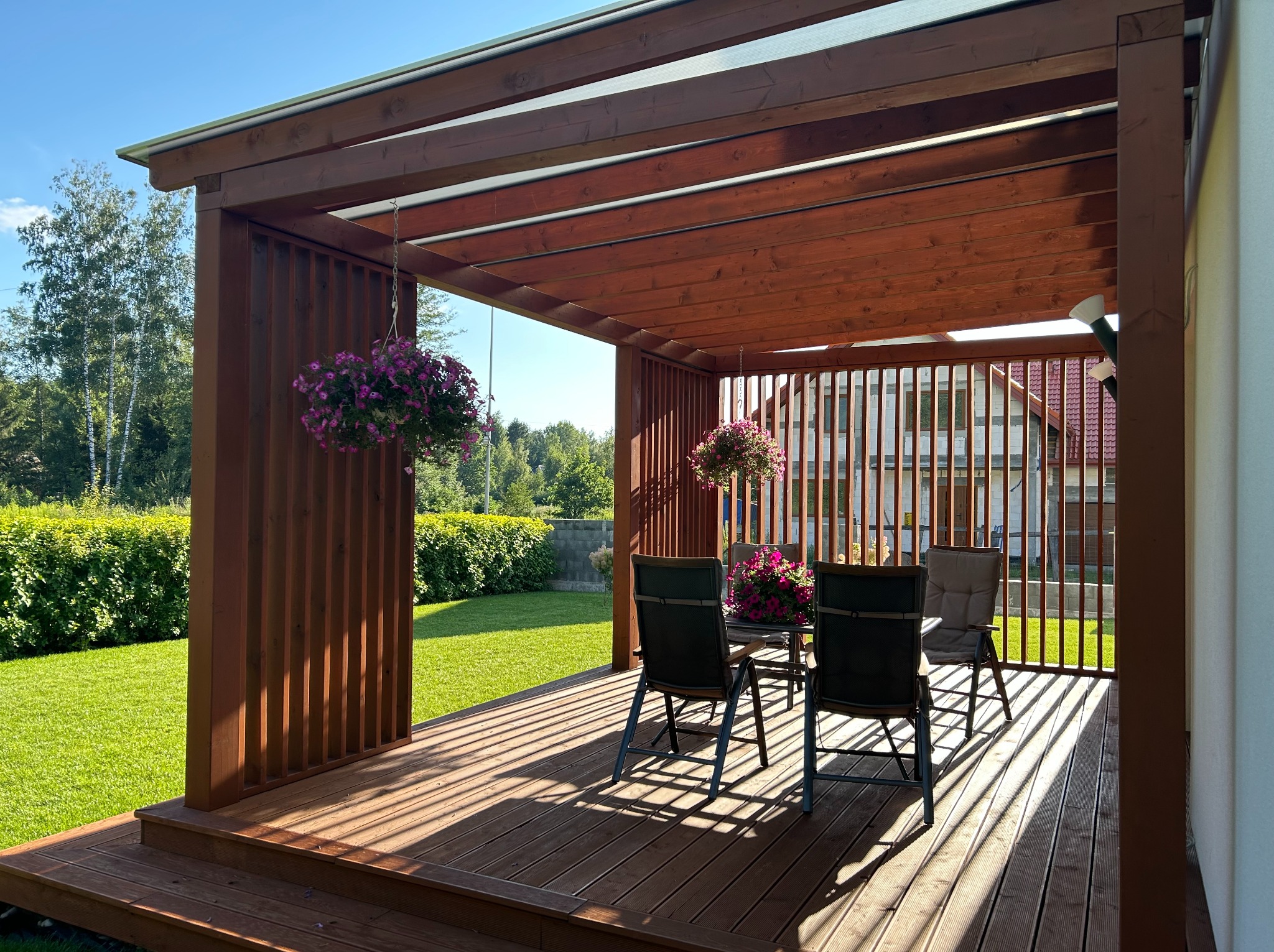 Drewniana pergola z ażurowymi ściankami bocznymi, zdobiona wiszącymi kwiatami, oświetlona promieniami słońca padającymi na taras z desek. Widok na zadbany ogród.