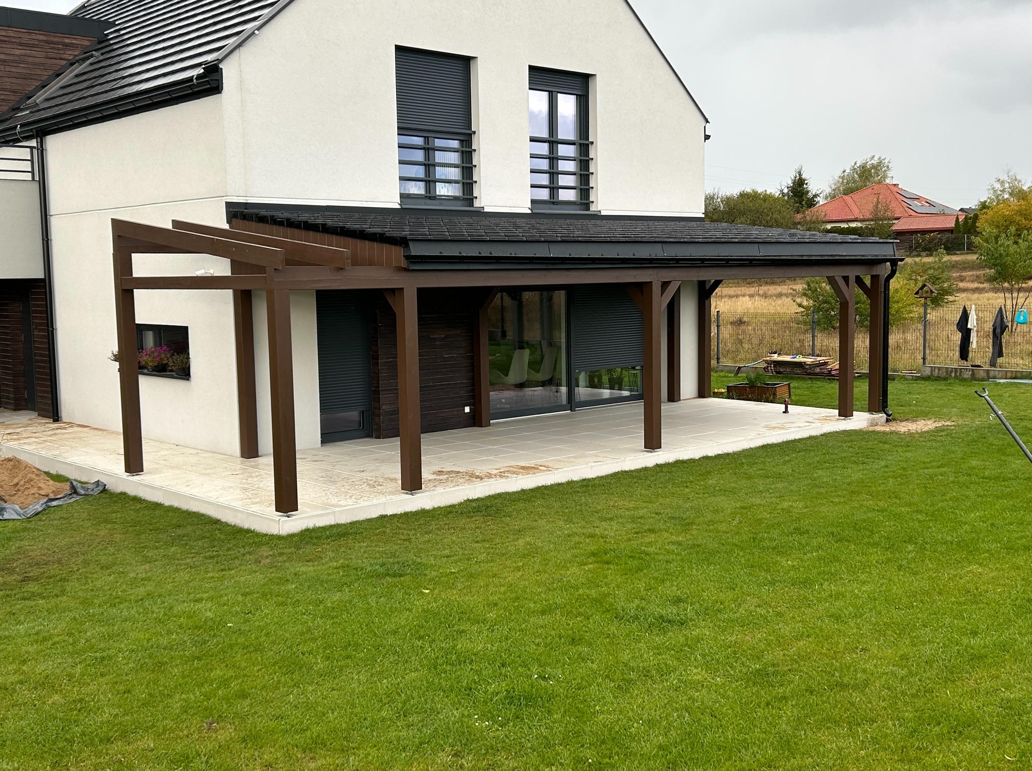 Nowoczesna, brązowa konstrukcja zadaszenia tarasu przy białym domu z ciemnymi roletami, otoczona zielonym trawnikiem. Widok na ogród i sąsiedni dom.