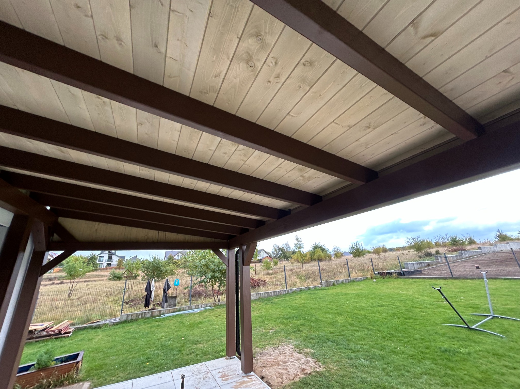 Widok z dołu na drewnianą konstrukcję pergoli z brązowymi belkami i jasnym drewnianym dachem, z widokiem na zielony trawnik i ogrodzenie w tle.