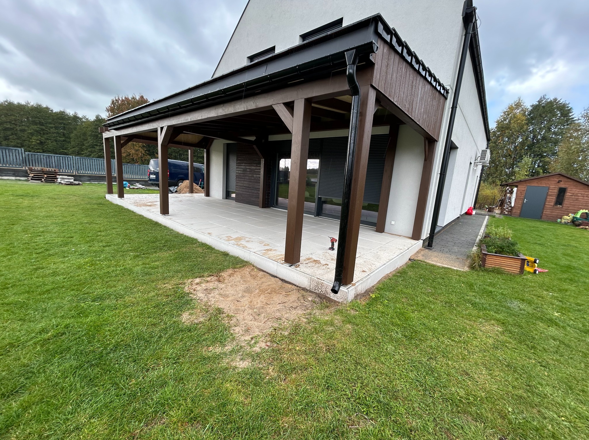 Nowoczesna pergola z ciemnego drewna przy białym domu z szarym tarasem, widok z trawnika, rynna, okna z żaluzjami, częściowo widoczny samochód w tle.