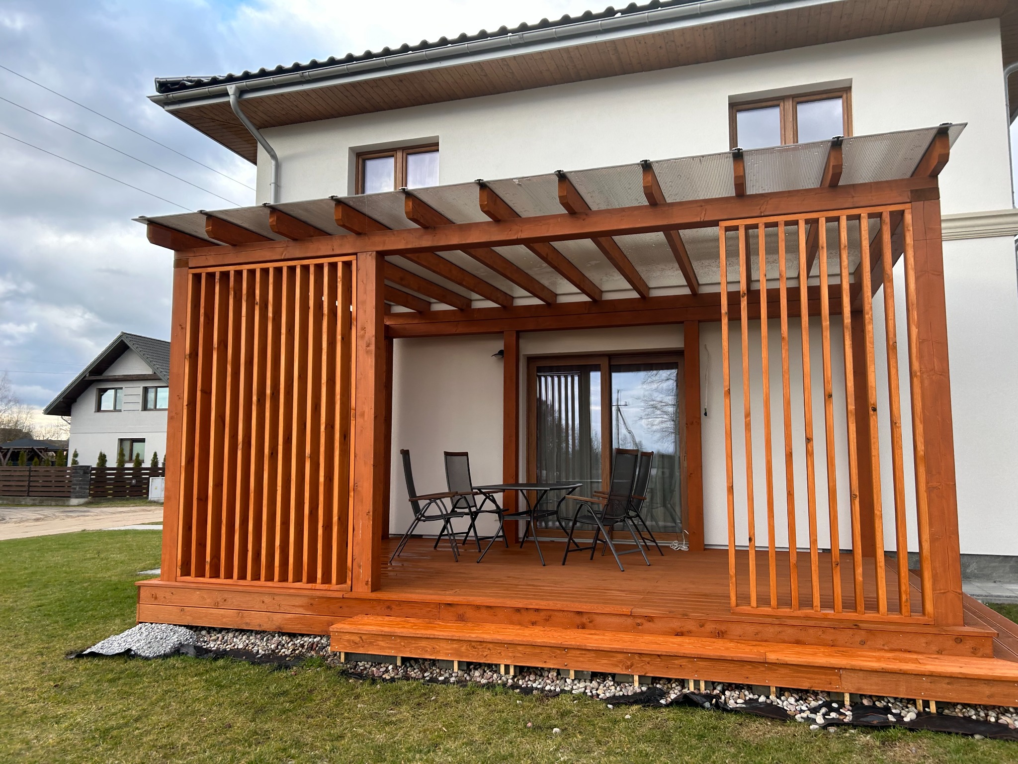 Drewniana konstrukcja z zadaszeniem przy domu. Na podeście stół z krzesłami. Pionowe deski tworzą ażurowe ściany. Widoczny trawnik i fragment domu.