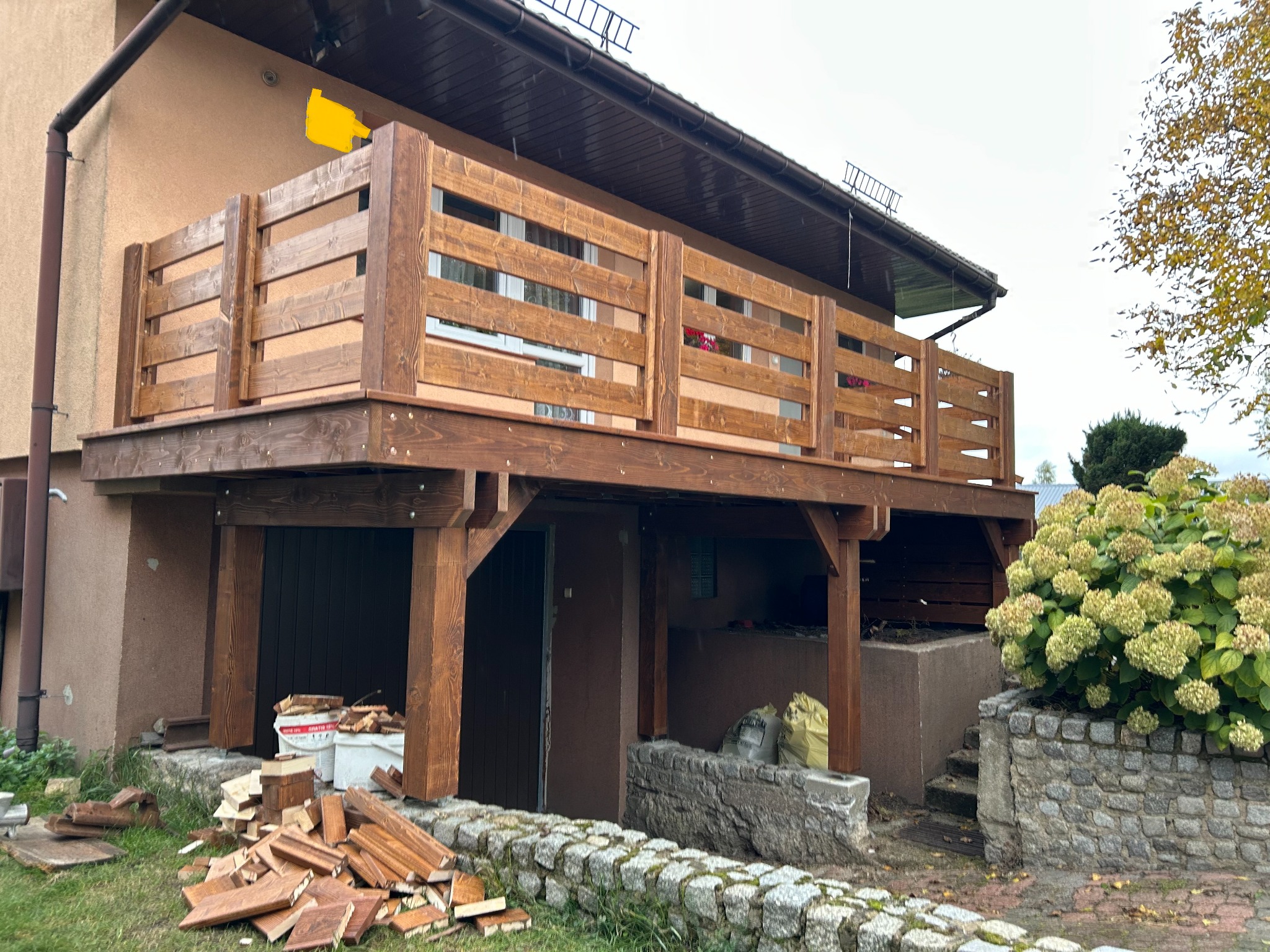 Drewniany balkon z balustradą w stylu rustykalnym, widok z dołu, z elementami ogrodu i resztkami materiałów budowlanych na trawie.