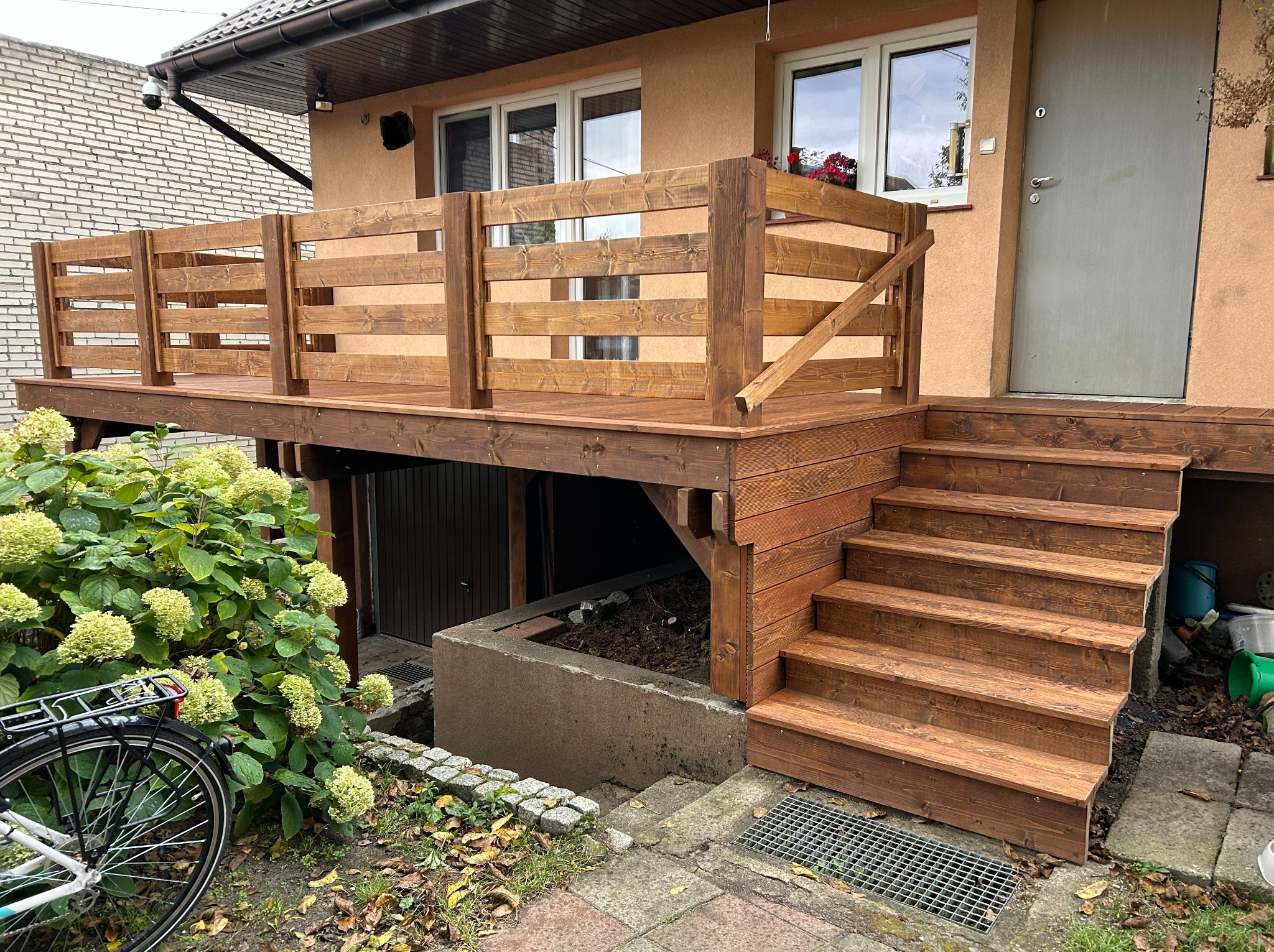 Drewniany taras z balustradą i schodami przy domu, z widocznym rowerem i hortensjami. Taras w odcieniach brązu, z wejściem do budynku. Widok na elewację.