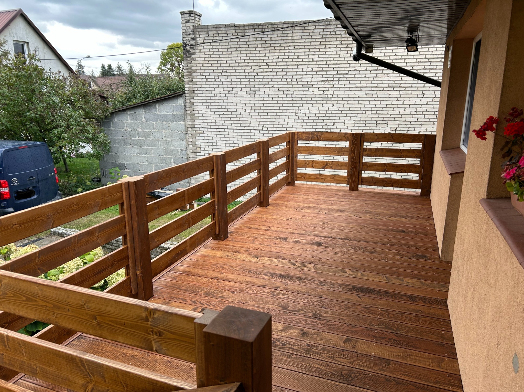 Nowy taras z drewna z balustradą, widok z góry, na tle budynku z cegły i fragmentu auta. Widoczne kwiaty w doniczce przy ścianie budynku.