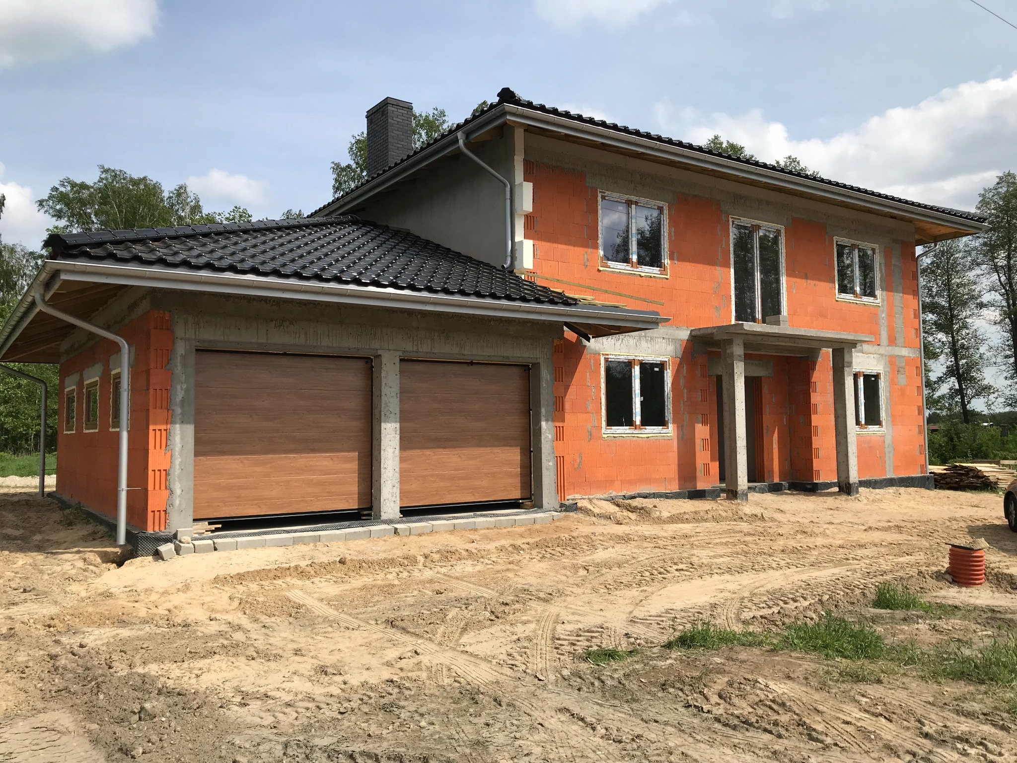 Dwukondygnacyjny dom w trakcie budowy, z garażem z brązowymi bramami, widoczne bloczki ceramiczne i szary dach z kominem, teren wokół w stanie surowym.