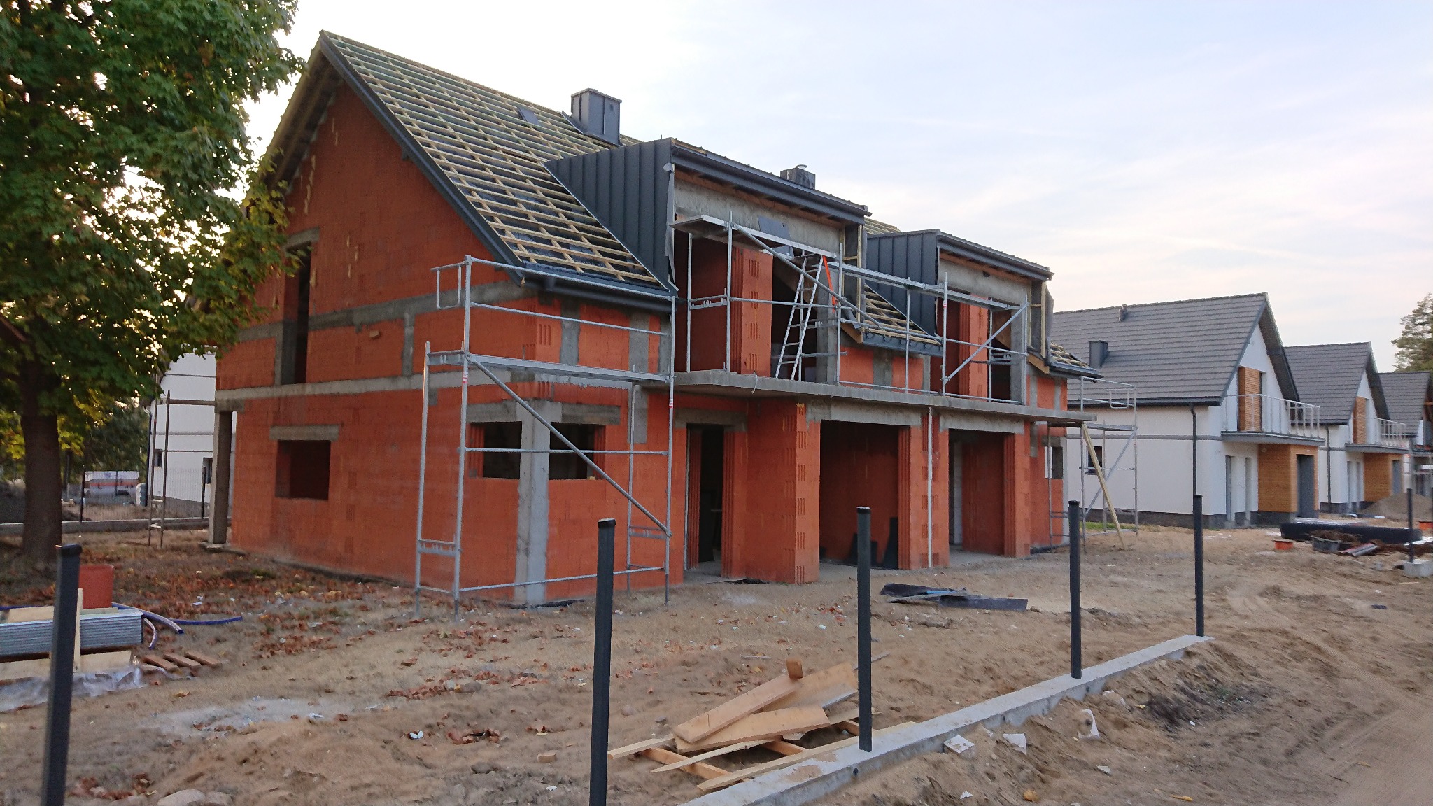 Budowa szeregówki z czerwonej cegły, widoczne rusztowania i elementy konstrukcji dachu. W tle gotowe domy z szarymi dachami i białymi elewacjami.