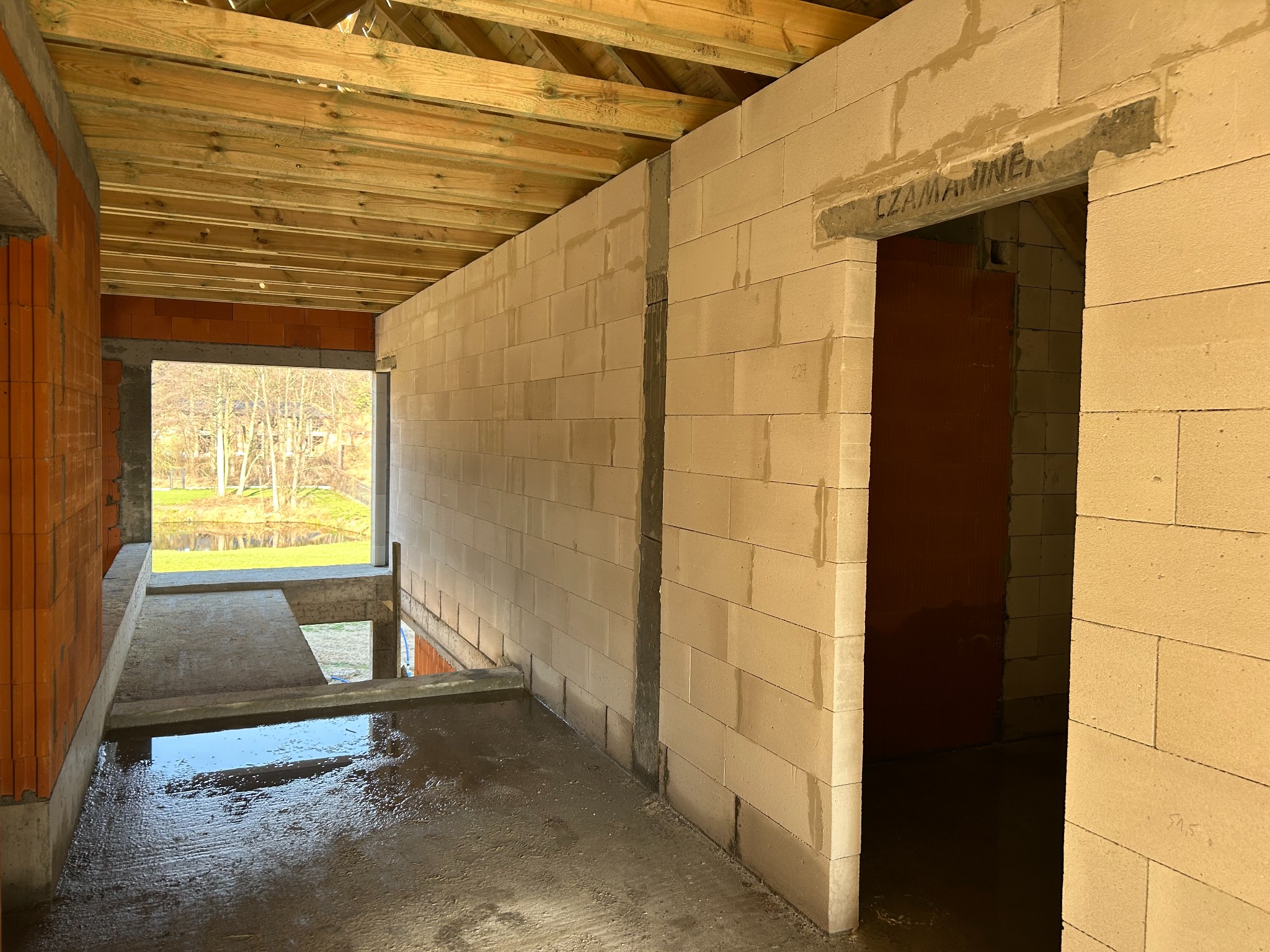 Wnętrze surowego domu w trakcie budowy: ściany z bloczków, drewniany strop, wylewka podłogowa z kałużami. Widok na zewnątrz przez otwór okienny.