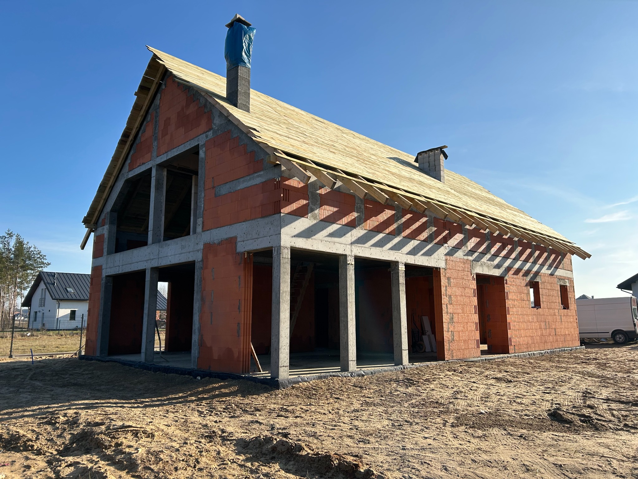Surowy stan domu z czerwonej cegły i betonu, z drewnianą konstrukcją dachu w trakcie budowy, na tle błękitnego nieba i piaszczystego terenu.
