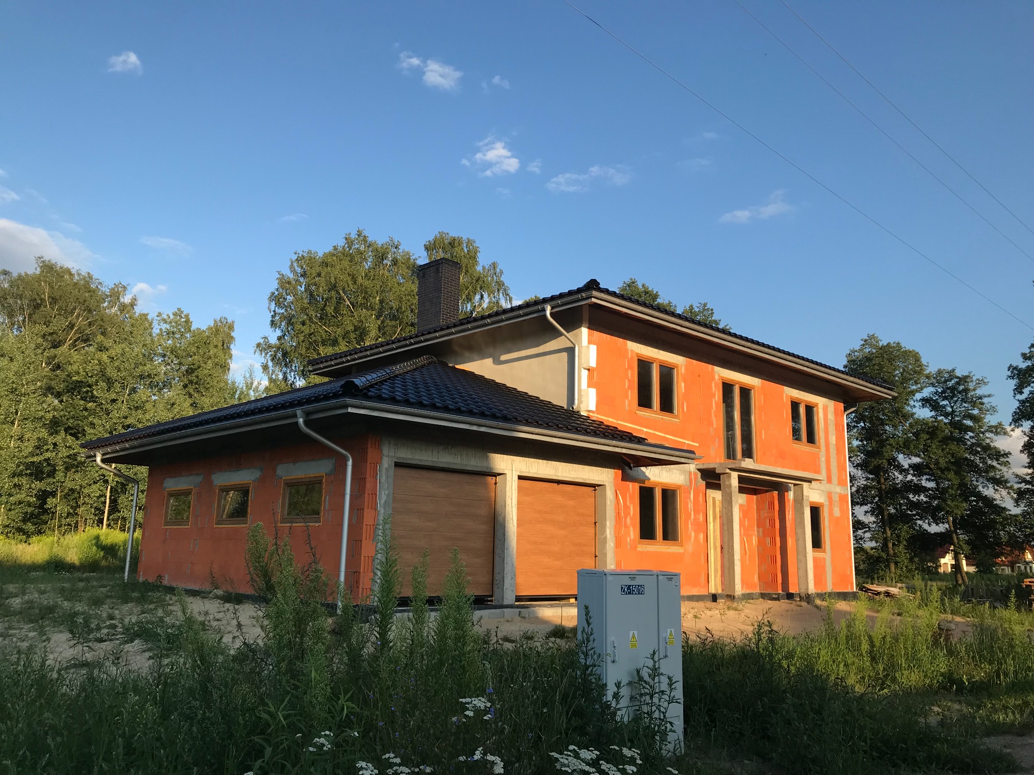 Dwukondygnacyjny dom w stanie surowym otwartym z garażem, zbudowany z czerwonej cegły, z widocznymi oknami i drzwiami, na tle zieleni i błękitnego nieba.