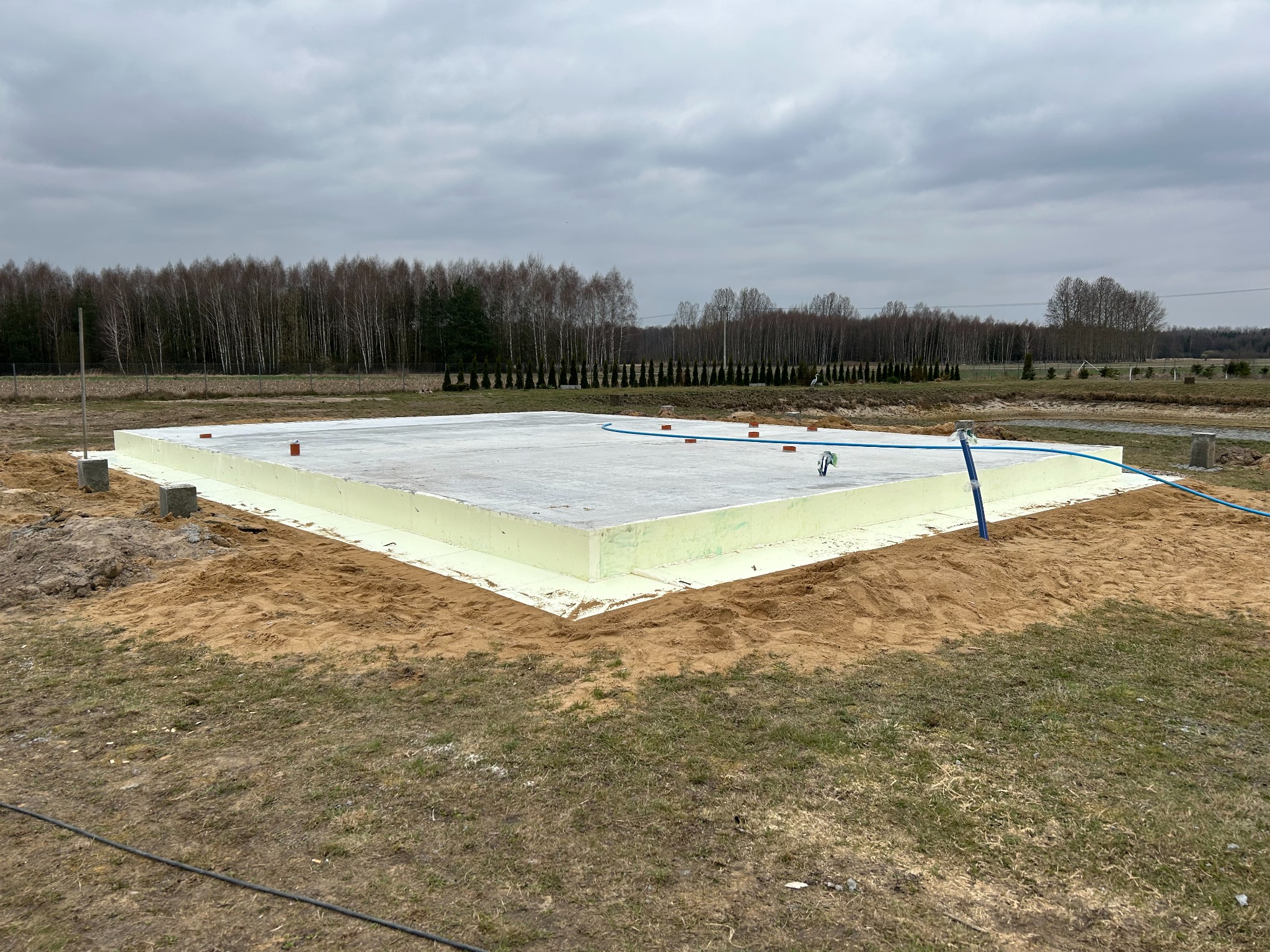 Płyta fundamentowa z izolacją termiczną, przygotowana do dalszych prac budowlanych. Widoczne cegły i niebieski wąż. Tło stanowią drzewa i pochmurne niebo.