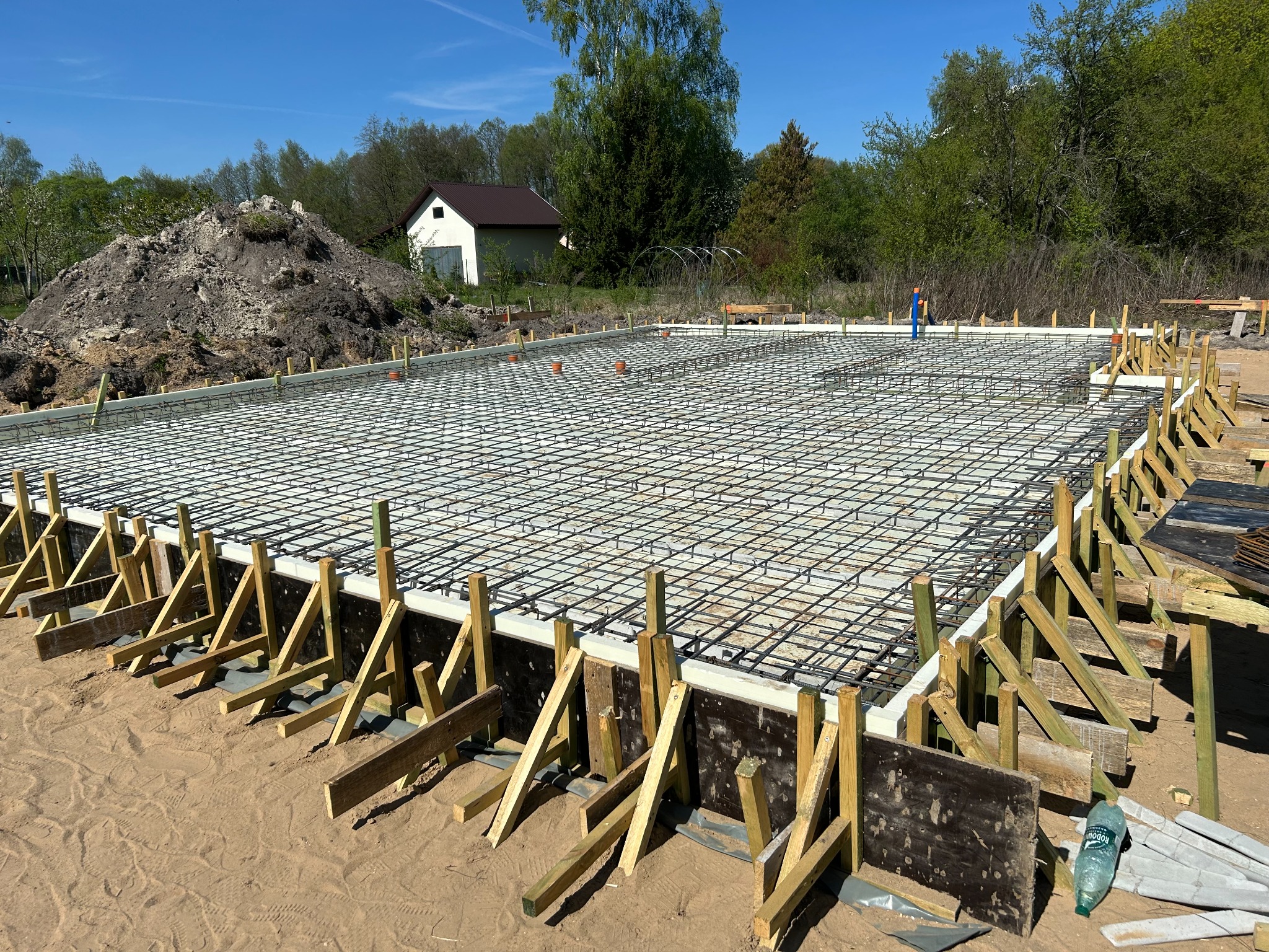 Zbrojenie fundamentu pod budowę domu jednorodzinnego. Widoczne szalunki, stalowe pręty zbrojeniowe, izolacja termiczna i drewniane podpory. W tle dom i drzewa.