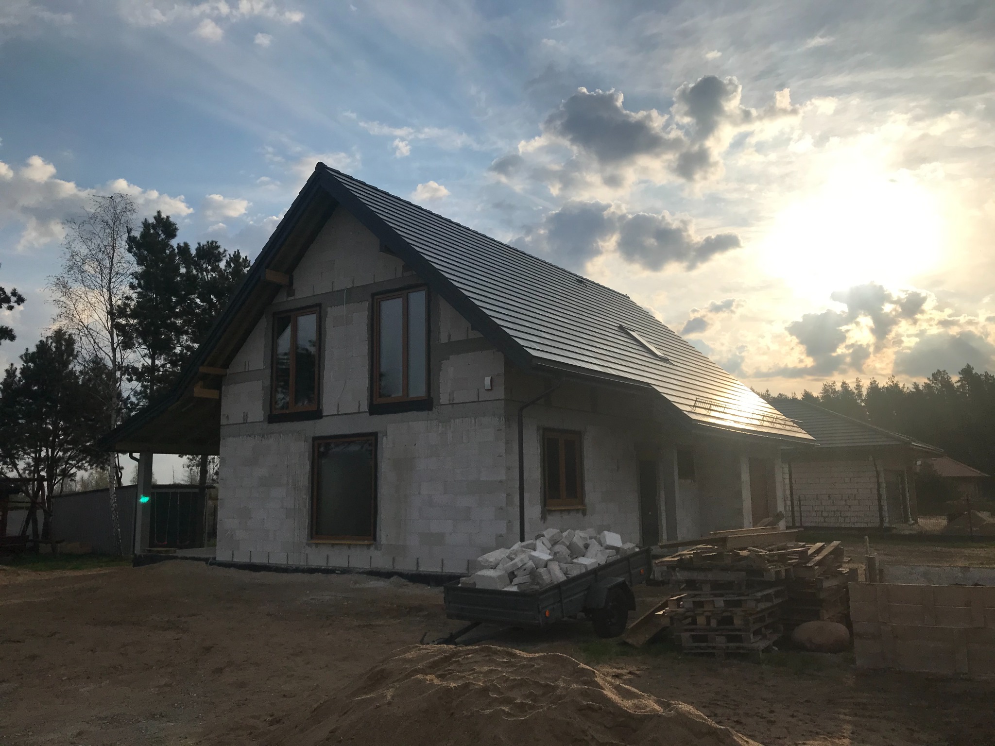 Surowy dom z bloczków, z ciemną dachówką, w trakcie budowy. Widoczne okna, przyczepka z materiałem i palety. Słońce za chmurami.