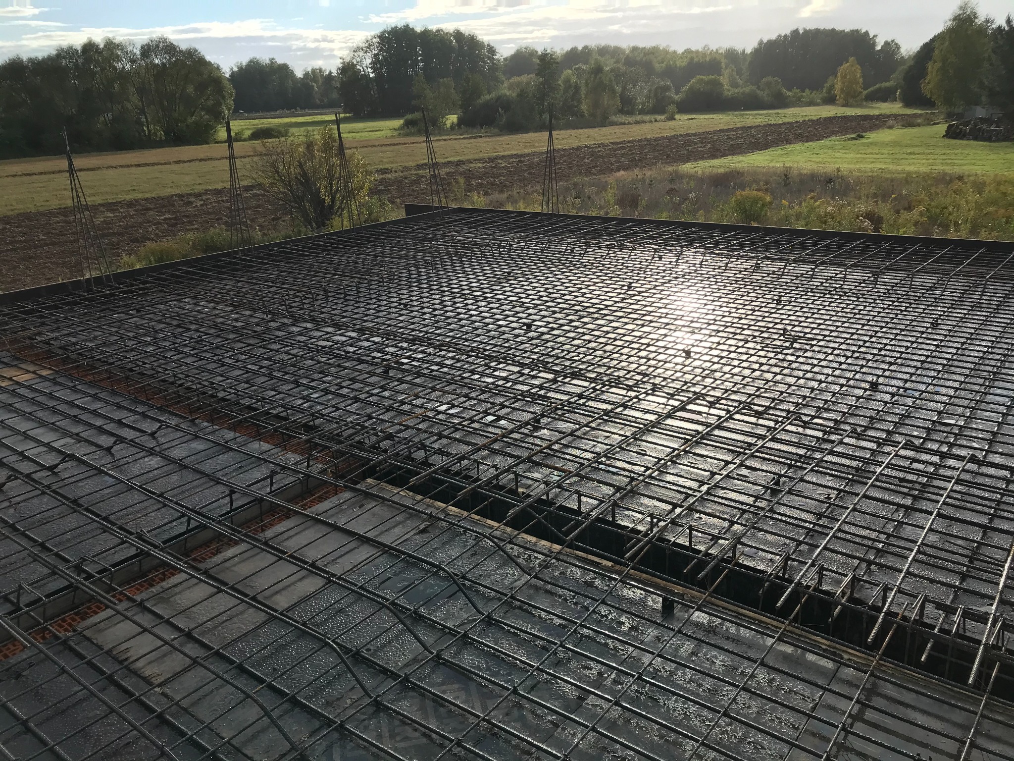 Zbrojenie płyty fundamentowej z widoczną siatką stalową i wystającymi prętami, przygotowane do zalania betonem na tle krajobrazu polnego.