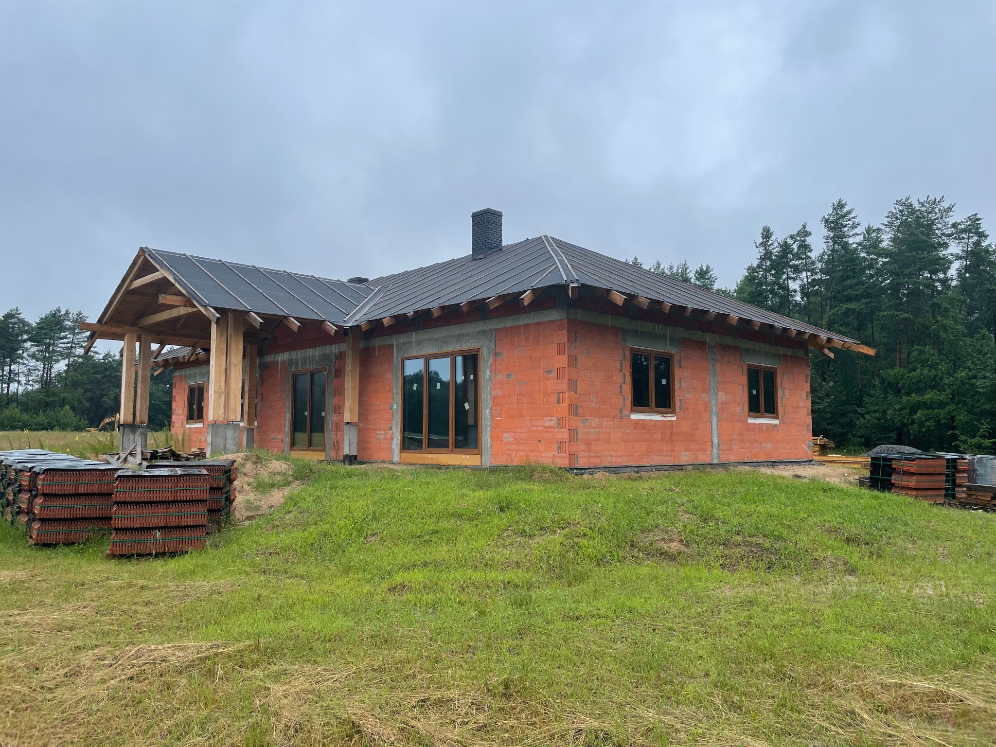 Niedokończony dom murowany z czerwonej cegły z ciemnym dachem i drewnianą konstrukcją werandy, otoczony zielonym trawnikiem i lasem w tle.