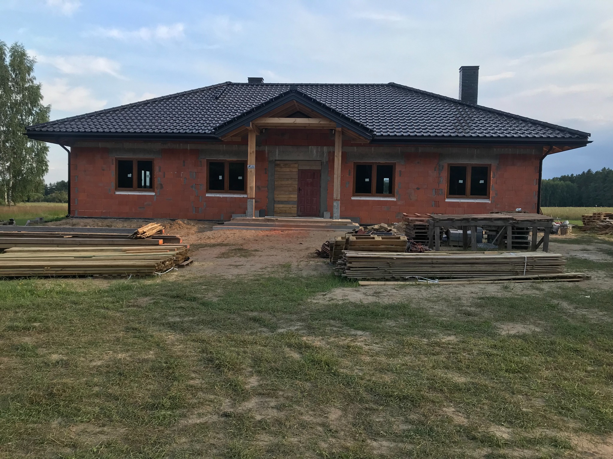 Niedokończony dom z czerwonej cegły z ciemnym dachem i drewnianą konstrukcją ganku. Na trawie leżą deski. Widoczne okna i drzwi zabite deskami.