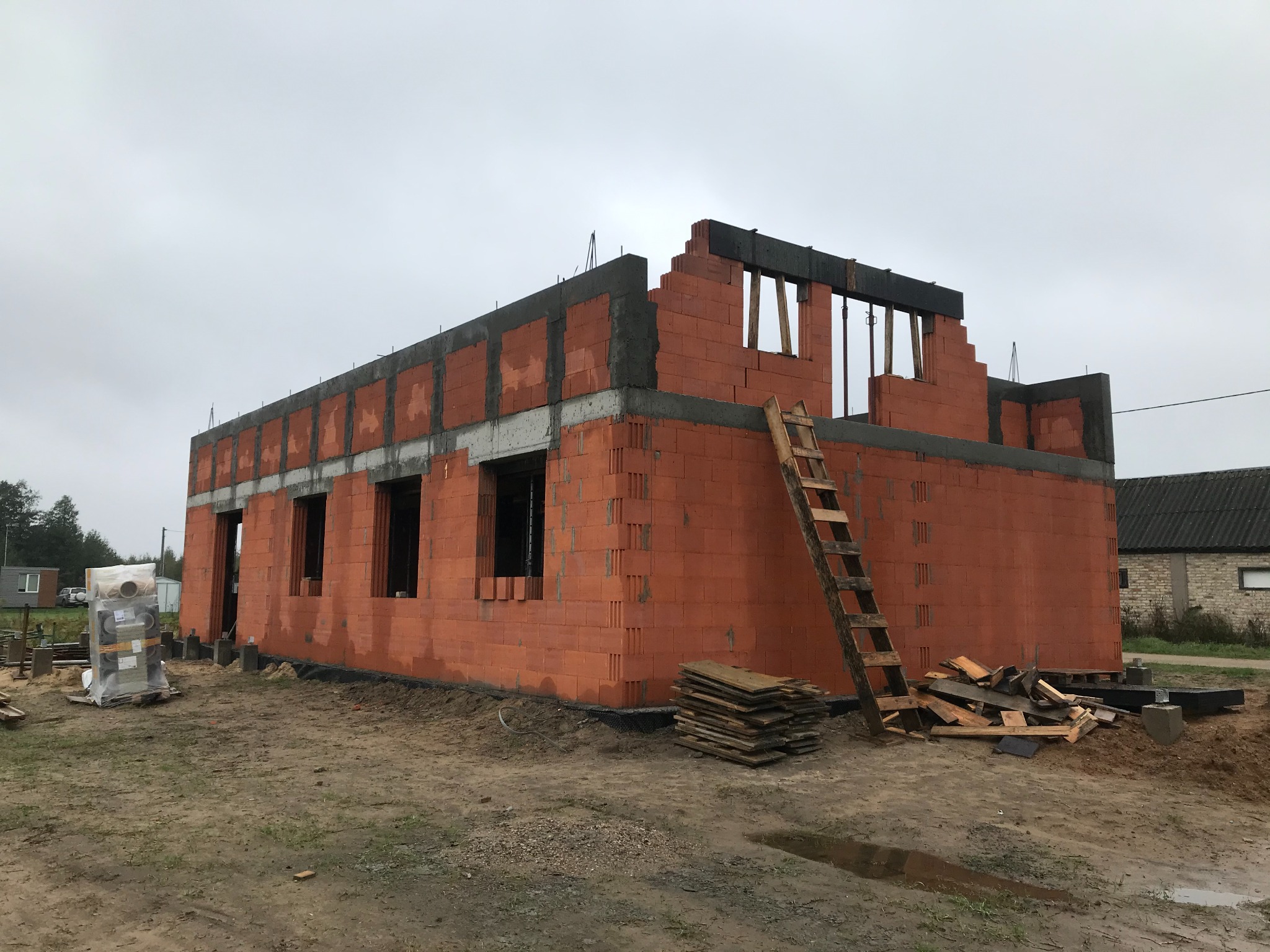 Budowa domu z czerwonej cegły ceramicznej, widoczne ściany parteru i fragment pierwszego piętra, drabina oparta o ścianę, materiały budowlane obok.