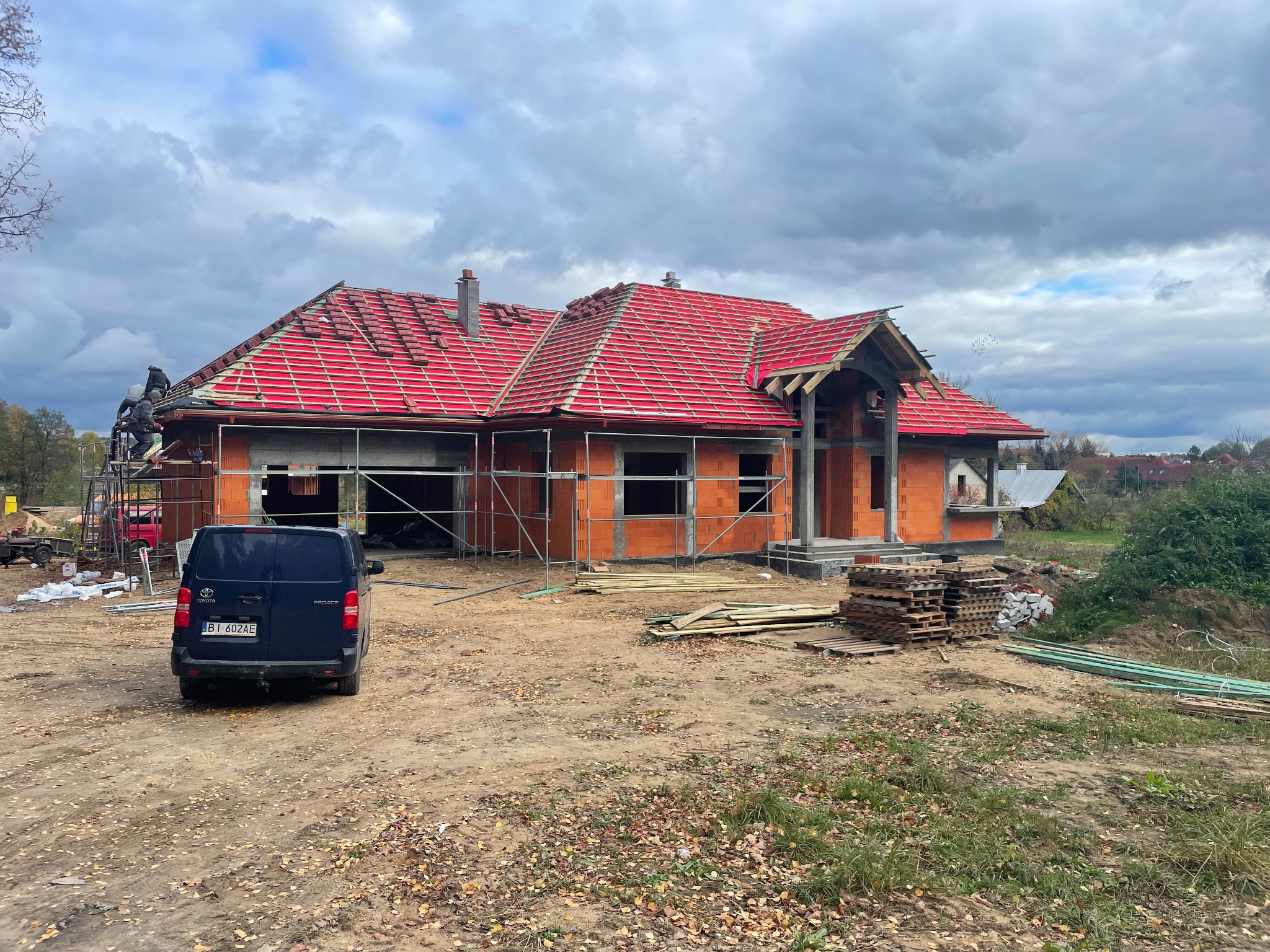 Surowy stan domu z czerwonej cegły z częściowo ułożonym czerwonym dachem, rusztowaniami i zaparkowanym vanem na placu budowy w pochmurny dzień.