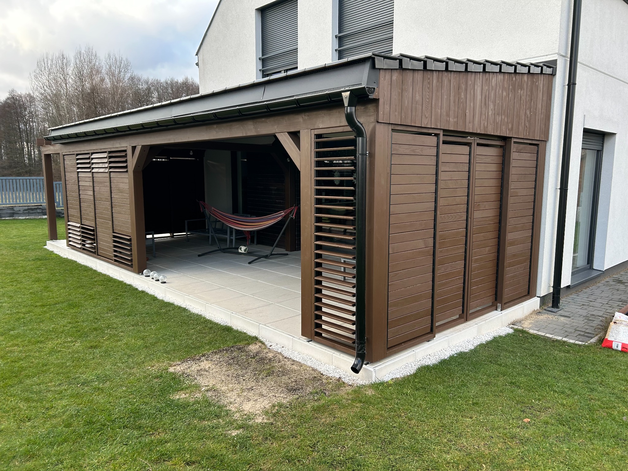 Nowoczesna pergola z brązowego drewna z lamelami i hamakiem w środku, przylegająca do białego domu z szarymi roletami, otoczona zielonym trawnikiem.