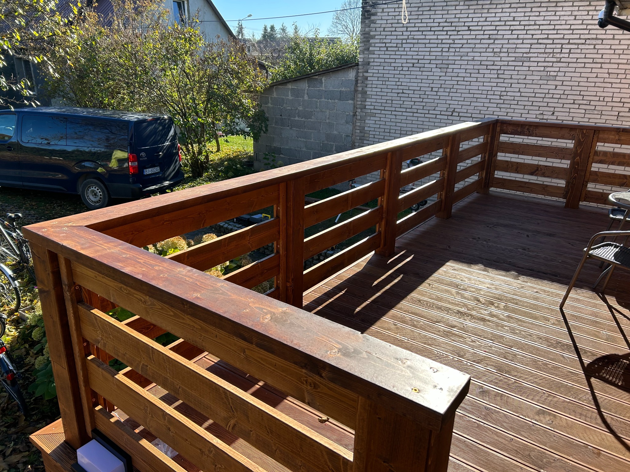Drewniany taras z balustradą w ciepłym odcieniu brązu, z widocznym fragmentem roweru i samochodu w tle, oświetlony naturalnym światłem słonecznym.