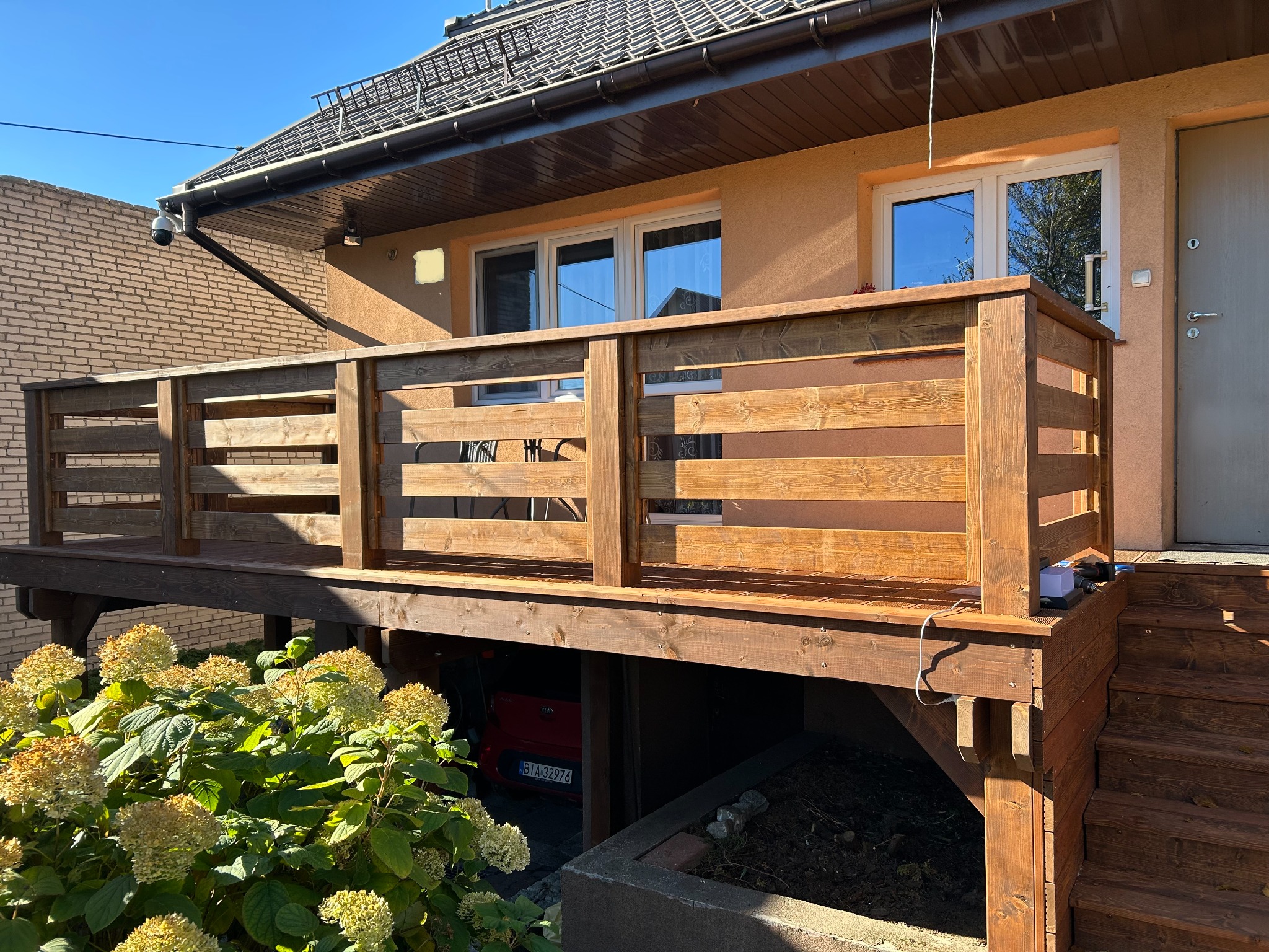 Drewniany taras z balustradą, widok z dołu na tle domu z beżową elewacją i brązowym dachem. Pod tarasem widoczny samochód. Krzewy hortensji w lewym dolnym rogu.