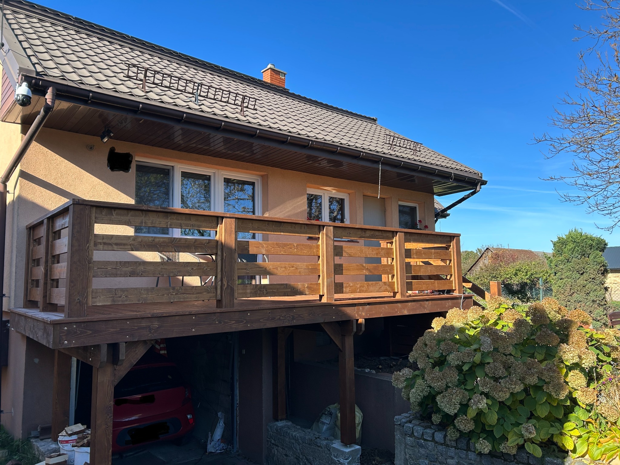 Drewniany taras z balustradą przy domu jednorodzinnym, wsparty na słupach nad garażem. Widok na elewację budynku i błękitne niebo w słoneczny dzień.