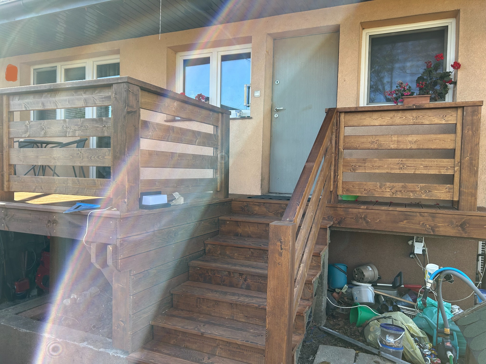 Drewniany taras z balustradą i schodami, wykończony w ciepłym brązie. Widoczne detale konstrukcyjne i naturalne usłojenie drewna. W tle elewacja budynku z oknami i drzwiami.