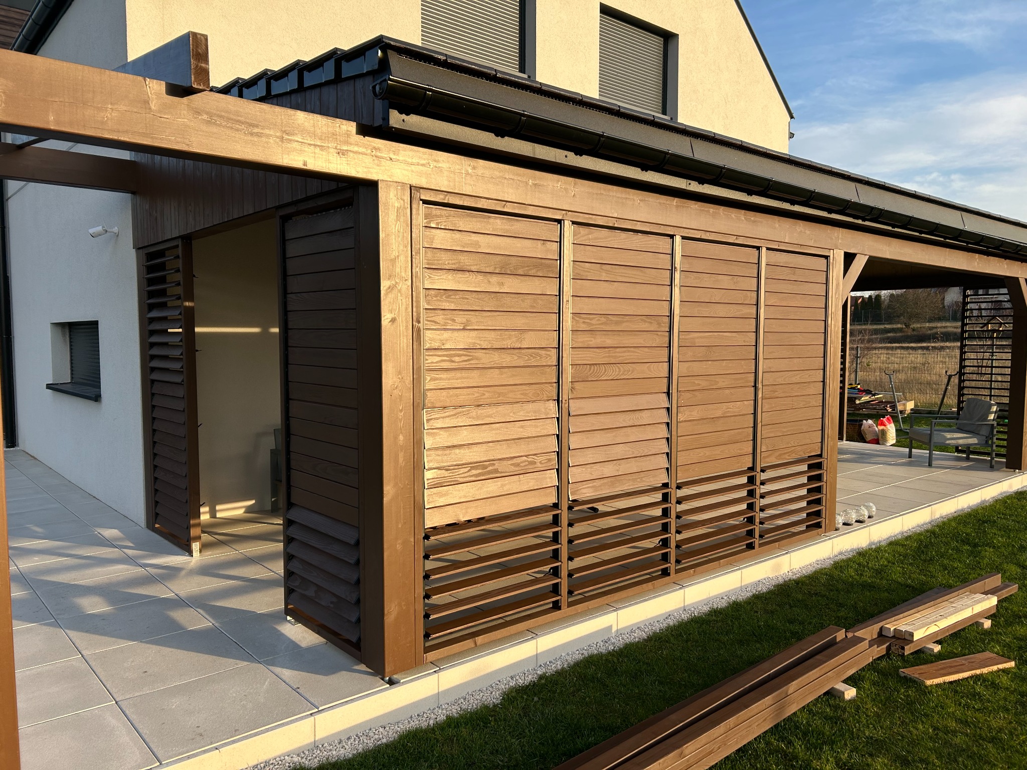 Nowoczesna pergola z drewnianymi żaluzjami w kolorze brązowym, przylegająca do jasnego budynku. Widoczne elementy konstrukcyjne i fragmenty tarasu z płyt.