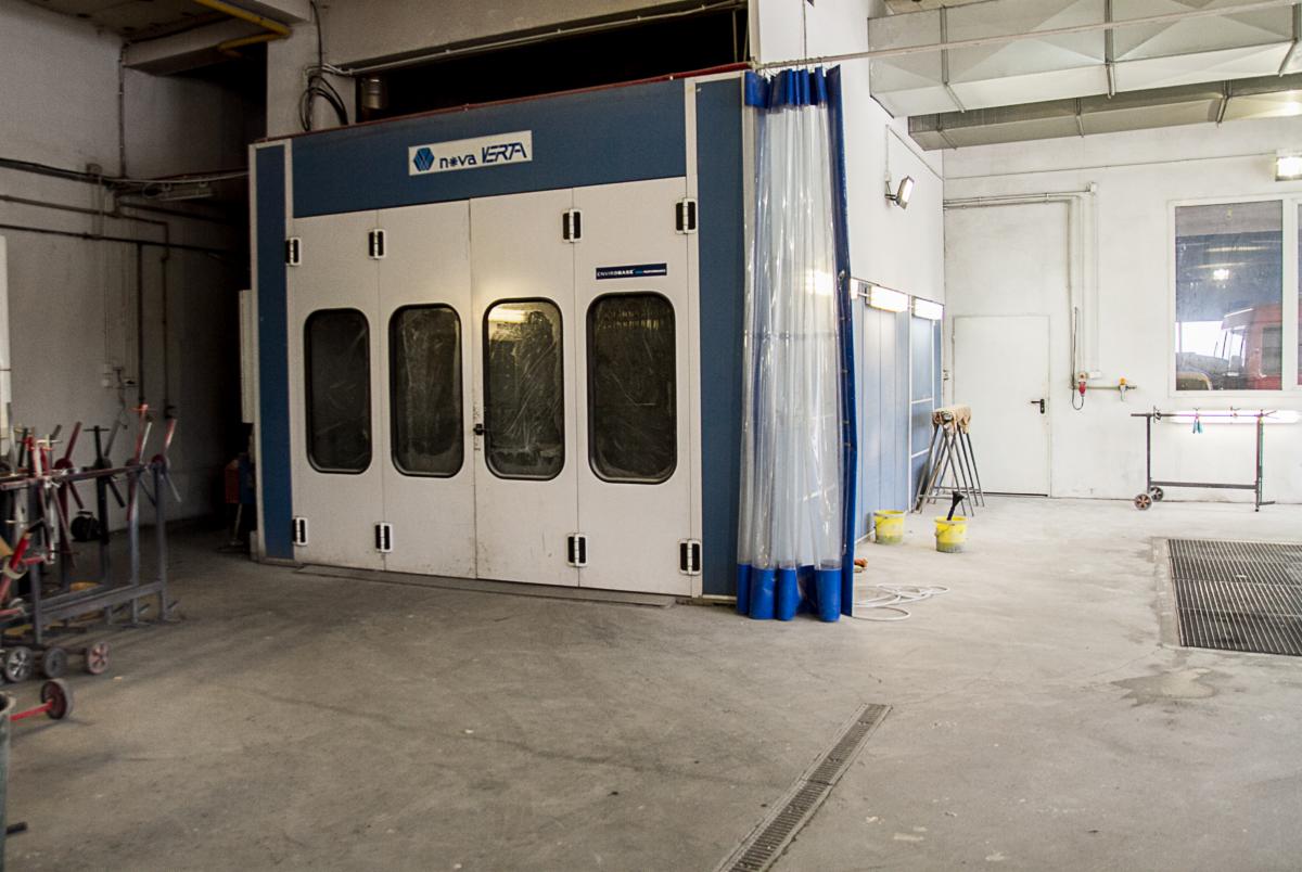 Wnętrze warsztatu samochodowego z kabiną lakierniczą Nova Verta, widoczne wózki narzędziowe oraz żółte wiadra, posadzka betonowa z kratką odpływową.