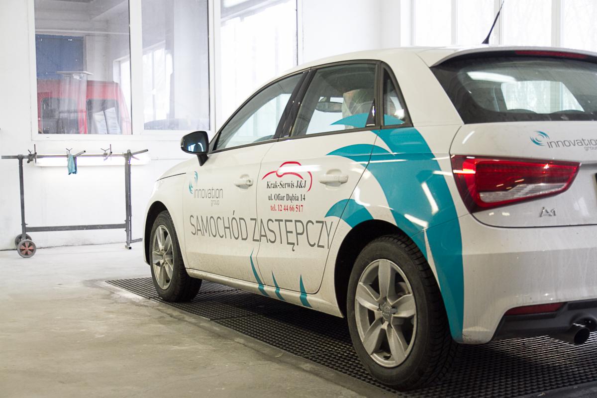 Białe Audi A1 z niebieskimi grafikami i napisem 'Samochód Zastępczy' zaparkowane w warsztacie, widoczne narzędzia i oświetlenie w tle.