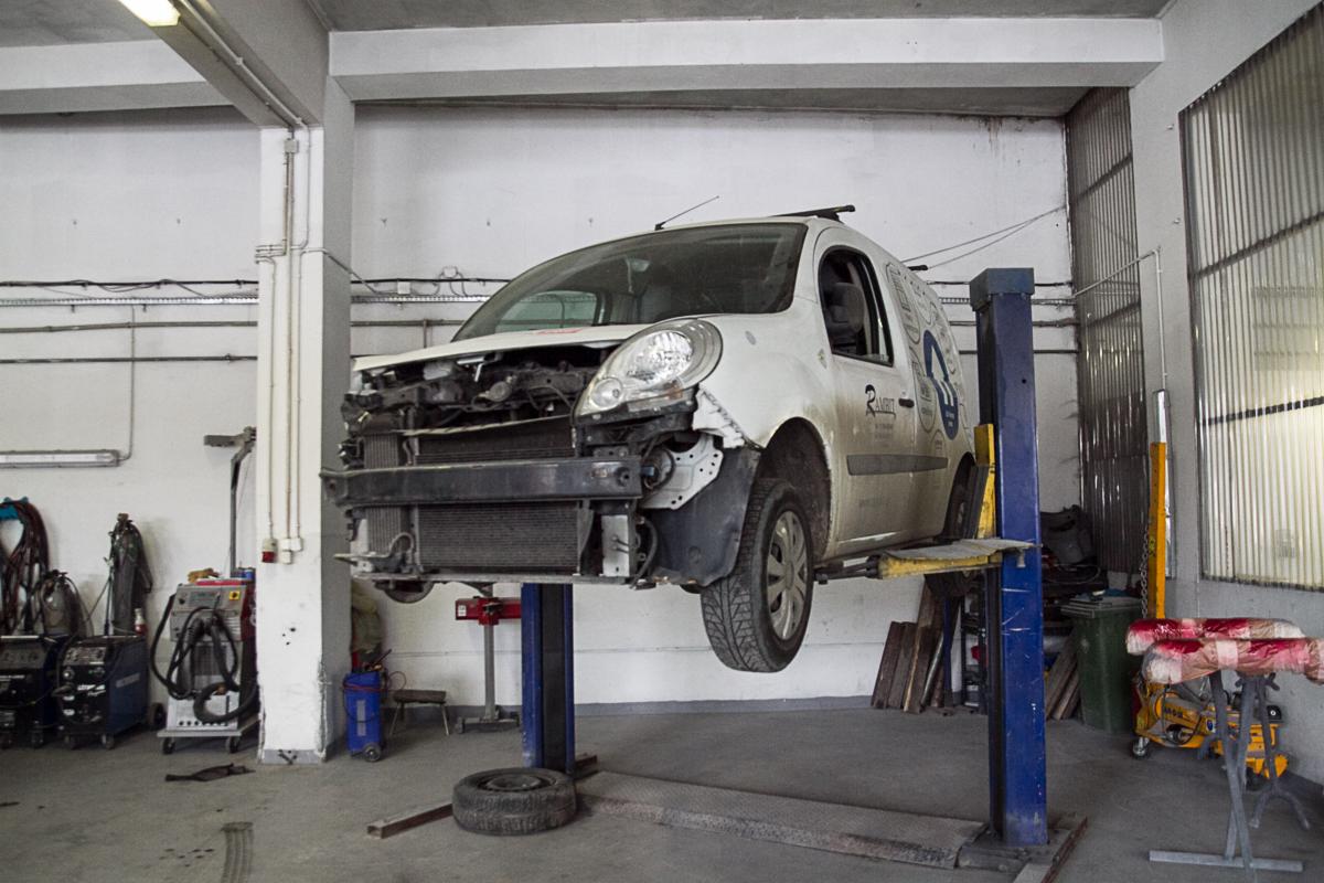 Uszkodzony biały samochód dostawczy na podnośniku w warsztacie w Krakowie, widoczne uszkodzenia przedniego zderzaka i brak elementów karoserii, w tle sprzęt warsztatowy i oświetlenie jarzeniowe.