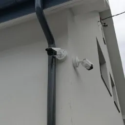 Dwie białe kamery monitoringu zamontowane na rogu białego budynku, jedna przymocowana do rynny, druga do ściany, widoczne przewody i pochmurne niebo w tle.