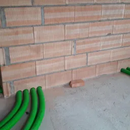 Wewnętrzne ściany z cegły ceramicznej z bruzdami, w których ułożono zielone rury karbowane, prawdopodobnie do rozprowadzenia instalacji wentylacyjnej lub rekuperacji.