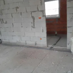 Surowy stan wnętrza budynku w trakcie prac instalacyjnych: widoczne przewody instalacyjne ułożone na podłodze, ściany z betonu komórkowego z widocznymi miejscami na puszki elektryczne, w tle ściana...