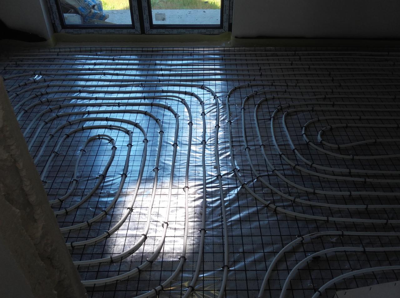 Białe rury ogrzewania podłogowego ułożone na czarnej siatce montażowej, widoczne okno i worek z materiałami budowlanymi w tle, wpadające światło słoneczne.