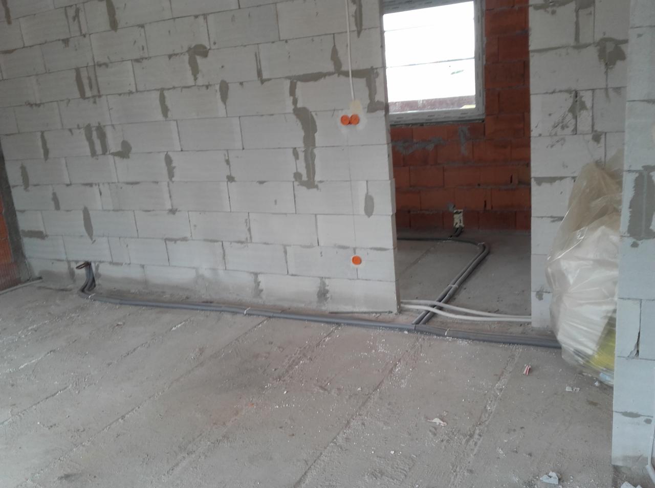 Surowy stan wnętrza budynku w trakcie prac instalacyjnych: widoczne przewody instalacyjne ułożone na podłodze, ściany z betonu komórkowego z widocznymi miejscami na puszki elektryczne, w tle ściana...