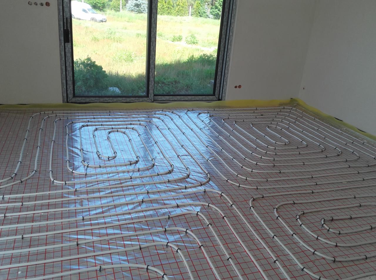 Białe rury systemu grzewczego ułożone w pętle na folii z czerwoną siatką, widok na okno z zielenią w tle, przygotowanie do wylewki.
