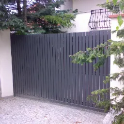 Nowoczesna, szara brama wjazdowa z pionowych paneli, otoczona zielenią i brukowaną nawierzchnią.