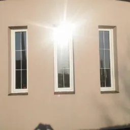 Elewacja budynku z czterema wąskimi, białymi oknami z podziałami, odbijającymi silne promienie słoneczne.