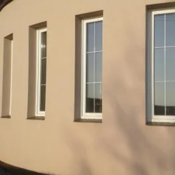 Fragment zaokrąglonej elewacji budynku w odcieniach beżu z trzema białymi oknami o różnej szerokości, z widocznym odbiciem krajobrazu i nieba w szybach, oświetlona popołudniowym słońcem rzucającym...