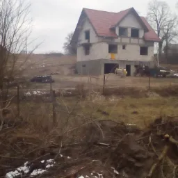 Surowy stan dwukondygnacyjnego domu z czerwonym dachem w trakcie budowy, widoczny mikser do betonu i traktor na zewnątrz, krajobraz wiejski w pochmurny dzień.