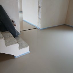 A&M Mariusz Batycki - Świeżo wylana, gładka wylewka anhydrytowa w jasnym pomieszczeniu, zabezpieczona niebieską taśmą przy ścianach i schodach. Widoczny fragment schodów i zwinięta folia.